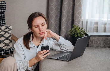 Focused woman uses her smartphone while sitting at a laptop, engaged in business communication or chatting.