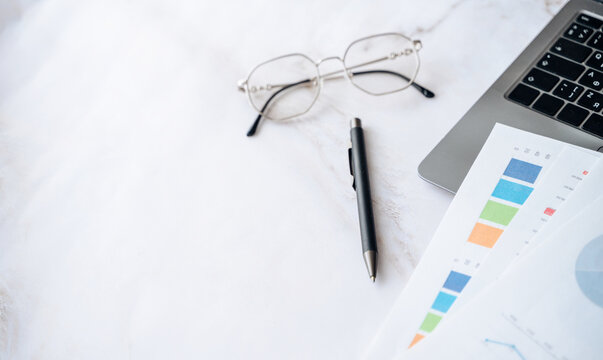 Desk setup with glasses, pen, laptop, and financial papers showing charts and graphs for business analysis