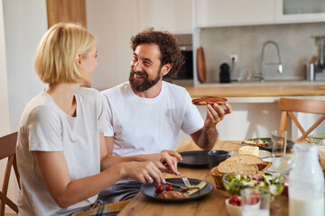 Two people are seated at a wooden table, enjoying a hearty breakfast. One is holding a slice of toast while the other cuts into a meal, surrounded by various dishes and drinks.