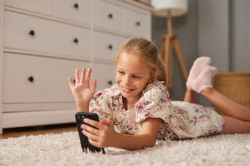 Fototapeta premium A girl lies on her stomach, smiling and waving at her phone during a video call in a warmly decorated bedroom filled with natural light.
