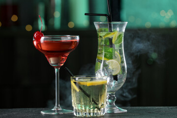 Glasses of tasty cocktails on dark table against blurred background with smoke, bokeh effect