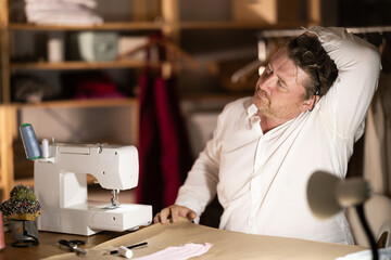 Middle-aged male tailor stretching while sitting next to sewing machine at night
