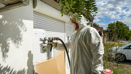 In meticulous harmony, a masked worker sprays vivacious white paint against the wall beneath leafy...