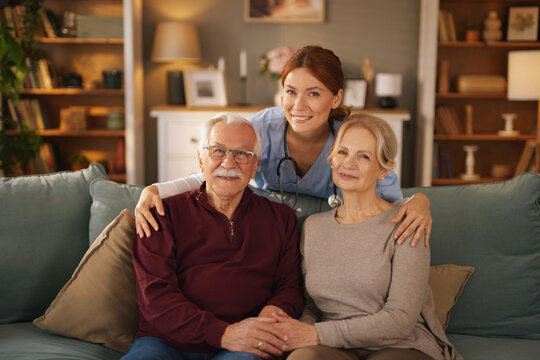A young female home caregiver smiles, posing with a senior couple in their home. The older man and woman are sitting on a green sofa. The caregiver has her arms around them. - Powered by Adobe
