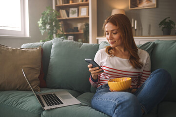 Obraz premium A woman sits comfortably on a couch in a cozy living room, using her smartphone while enjoying a bowl of popcorn. A laptop rests nearby, completing her relaxed afternoon vibe.