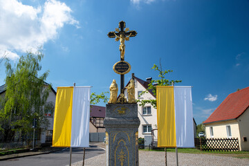 Vergoldetes Wegkreuz mit Kirchenfahnen in einem sorbischen Dorf der Lausitz. Katholische Osterdekoration als Zeichen des Glaubens und der Tradition in Sachsen