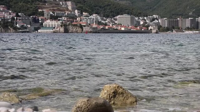 Sveti Nikola Island. Sea waves and stones. Old Town of Budva, Montenegro, in the background