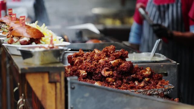 Vendor in gloves serves fried chicken and burger with white sauce at vibrant market stall, food in focus, background with blurred chef.