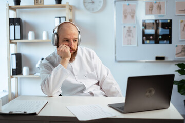 Tired lazy Bearded Male Doctor wearing white coat in Hospital Office using Laptop computer, Analyzing Medical Data.