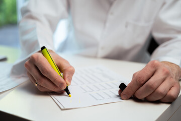 Close-up of male doctor's hands highlighting numbers on financial report with colored marker at desk in clinic