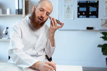 Bearded doctor using smartphone while listening voice message and working in medical clinic