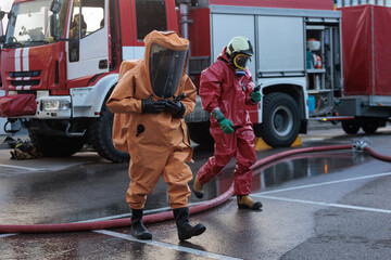 Two emergency responders in full chemical or biohazard suits and breathing apparatus walking in front of a red fire truck at a hazardous material incident scene.