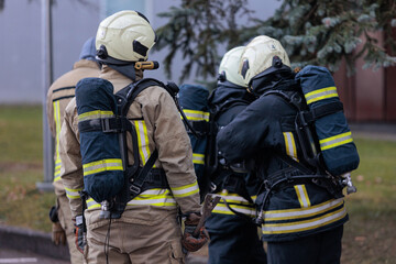 Obraz premium View from behind of a group of firefighters in protective suits, helmets, and oxygen tanks gathering for instruction or resting at an emergency scene.