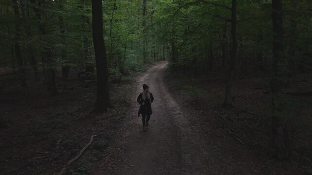 A person with a backpack walks along a narrow dirt path surrounded by tall trees and lush greenery in a peaceful forest. The natural setting evokes a sense of solitude and connection with nature.