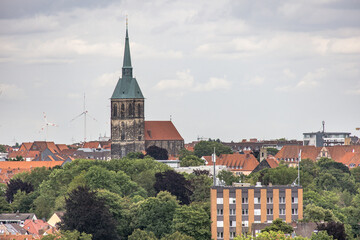Panoramic view of Hildesheim city with historic church tower, red rooftops, and green trees under cloudy summer sky