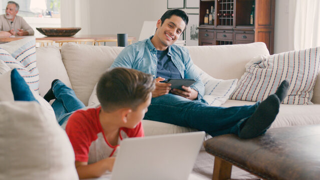 Father Sitting On Sofa At Home With Digital Tablet As Son Uses Laptop