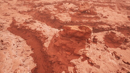A dramatic view of a vast, arid landscape dominated by red rocks and sandy soil. The rugged terrain features unique formations and shadows cast by the uneven surfaces, evoking a sense of isolation.