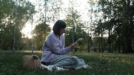 Young woman reading ebook sitting on grass in public park. Concept of recreating, resting, mindfulness, leisure without gadgets. Young woman is focused on reading or studying. - Powered by Adobe