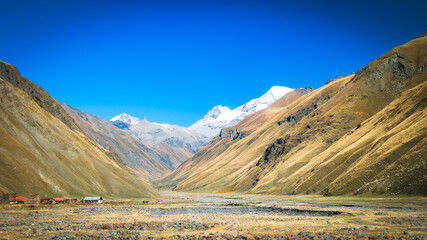Remote mountain valley near Truso Gorge in Georgia, with small rural houses, golden hills, and snowy peaks under a deep blue sky. Peaceful autumn vibes in the Caucasus