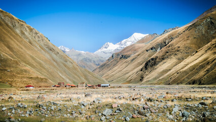 Remote mountain valley near Truso Gorge in Georgia, with small rural houses, golden hills, and snowy peaks under a deep blue sky. Peaceful autumn vibes in the Caucasus