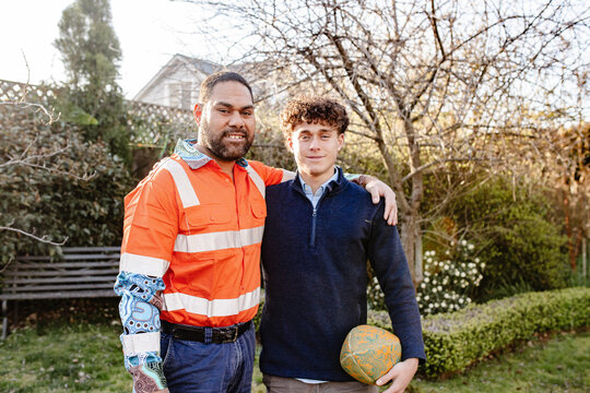 Fathers arm over teenage sons shoulder with one hand holding ruby ball standing in the garden