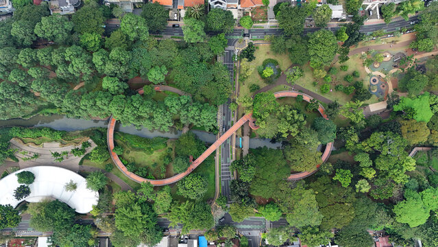 Aerial view of a vibrant orange infinity bridge winding through verdant trees and across a gentle river, Bogor, Jawa Barat, Indonesia.