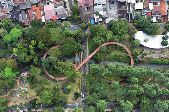 Aerial view of the vibrant, winding pedestrian bridge shaped like the infinity symbol cutting through a lush green park amidst urban dwellings, Jakarta, Indonesia.