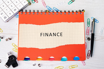 A notepad with the word FINANCE sits on a desk amid office supplies, a calculator, pen, flash drive, and paperclips. A concept for business and personal budgeting