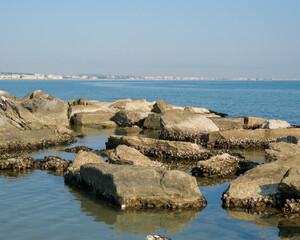 Low-angle view of a rugged, man-made rock breakwater in the calm sea. The modern skyline of the Italian resort town of Cattolica is visible in the background under a clear blue sky.