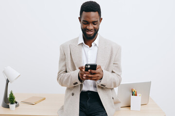 Smiling man with dark skin using smartphone in modern office setting, casual clothing, standing near desk with laptop, stationery, and potted plant, isolated on plain white background, studio shot