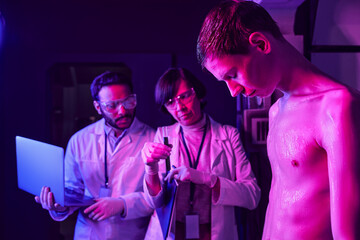 In a high tech lab, scientists closely examine a tall alien figure under vibrant purple lights