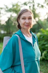 Happy active mature woman in blue sportswear with yoga mat on her shoulder outdoors. Sport training concept