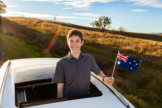 Aussie kid with Australian flag looking out sunroof of car parked beside rural road on ridge