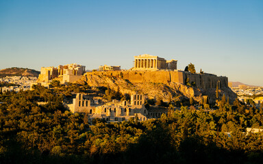 Fototapeta premium Acropolis of Athens during golden hour, with the Parthenon temple glowing above the city skyline. The soft evening light highlights the ancient ruins and the surrounding greenery, architecture