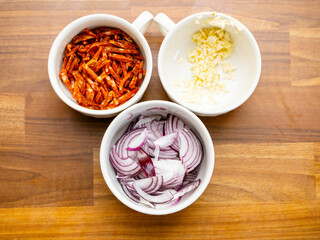 Three small cups with chopped onion, garlic and diced spicy chorizo sausage on wooden table. Pizza cooking ingredients. Food preparation.