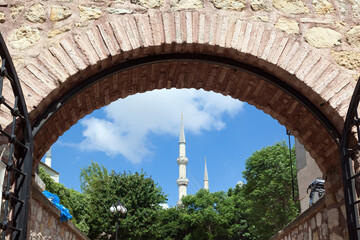 Beautiful view of famous Blue Mosque (Sultanahmet) minarets framed by an ancient stone archway in Istanbul, Turkey, under a bright blue sky with white clouds and green trees.