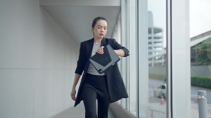 Asian businesswoman in smart casual outfit rushing through office hallway while checking time on smartwatch. Concept of urgency, pressure, and time management. - Powered by Adobe