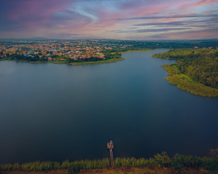 Aerial view of a serene lake reflecting the vibrant colors of the sunset sky, bordered by lush greenery and the distant cityscape, Iwo, Osun, Nigeria.