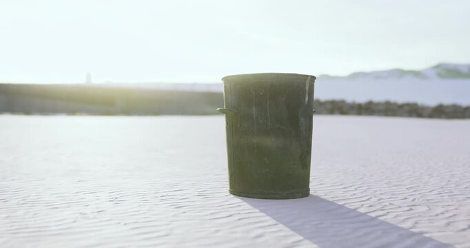 A dark bin rests amidst the sandy shoreline, illuminated by the gentle morning light. Waves softly lap at the coast, hinting at the tranquility of the scene, untouched except for this lone container.