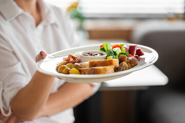 Person holding a white plate with assorted breakfast items: toast, jam, grilled mushrooms, cherry tomatoes, greens, and sausage. Casual dining setting, top view, natural light.