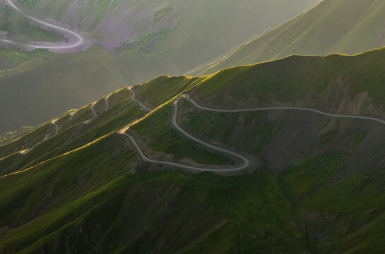 Aerial view of a serpentine road cutting through the rugged, verdant Alazani Pass, where sunlight paints the slopes with gold, Chala, Kakheti, Georgia.