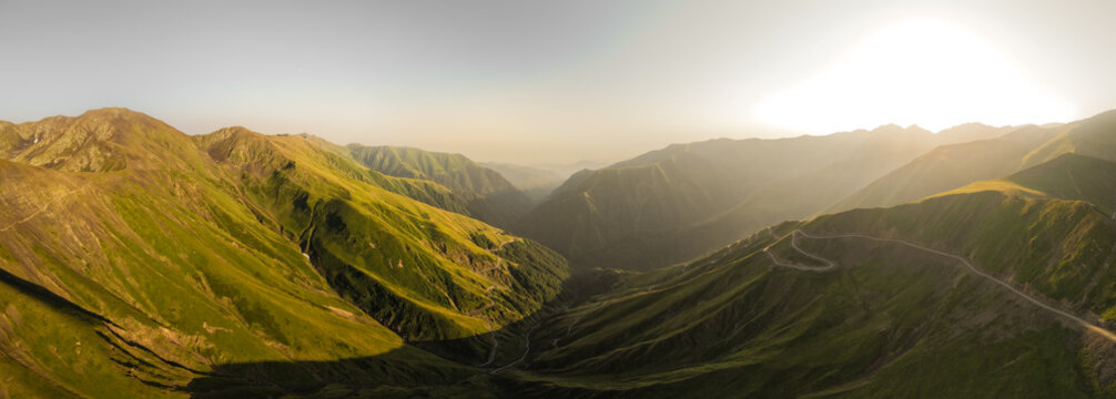 Aerial view of a winding road carving through the green, sun-kissed Alazani pass mountains under a hazy sky, Chala, Kakheti, Georgia.