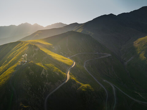 Aerial view of a winding mountain road bathed in golden sunlight cuts through verdant hillsides, leading to a distant peak, Chala, Kakheti, Georgia.