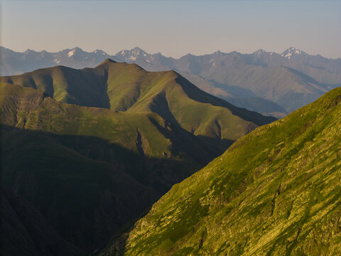 Aerial view of rolling green hills cascading into shadowy valleys with snow-capped peaks gracing the distant horizon, Chala, Kakheti, Georgia.