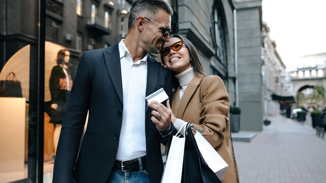 Stylish couple walking in city for shopping, smiling and hugging. Man in dark suit, woman in camel coat and sunglasses holding bags and credit card. Love and luxury lifestyle. Black friday sales - Powered by Adobe