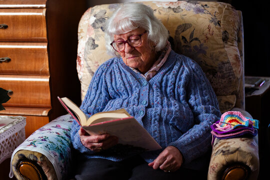 Older Aussie woman sitting on armchair in loungeroom reading a fiction novel