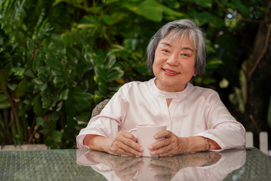 Portrait of a happy senior Asian woman smiling while holding a cup of coffee or tea in the garden. Enjoying a relaxed and healthy retirement lifestyle outdoors.
