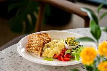 Healthy brunch plate with scrambled eggs, grilled toast, avocado, cherry tomatoes, and greens on a white dish. Natural light, cozy indoor setting, rustic and fresh presentation.