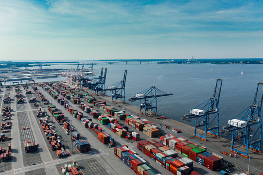 Aerial view of colorful containers stacked in neat rows at the port, with cranes reaching towards the sky against the backdrop of the Chesapeake Bay Bridge, Baltimore, Maryland, United States.