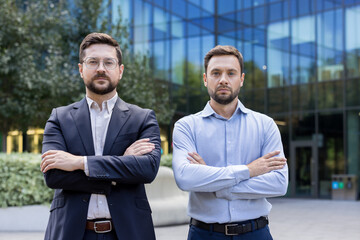 Two serious businessmen, standing with arms crossed in front of a modern glass office building,...